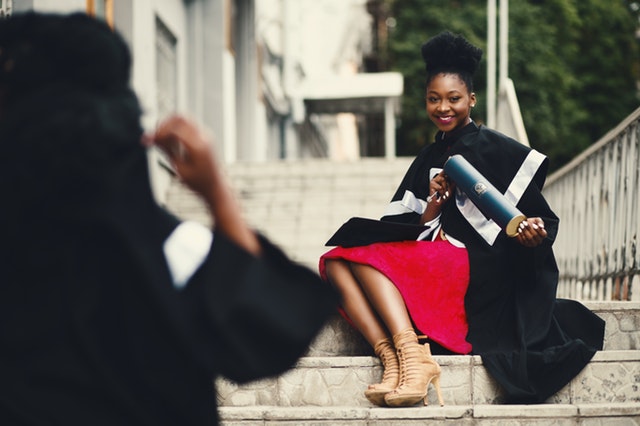 A woman in her graduation gown, holding her diploma and sitting on steps